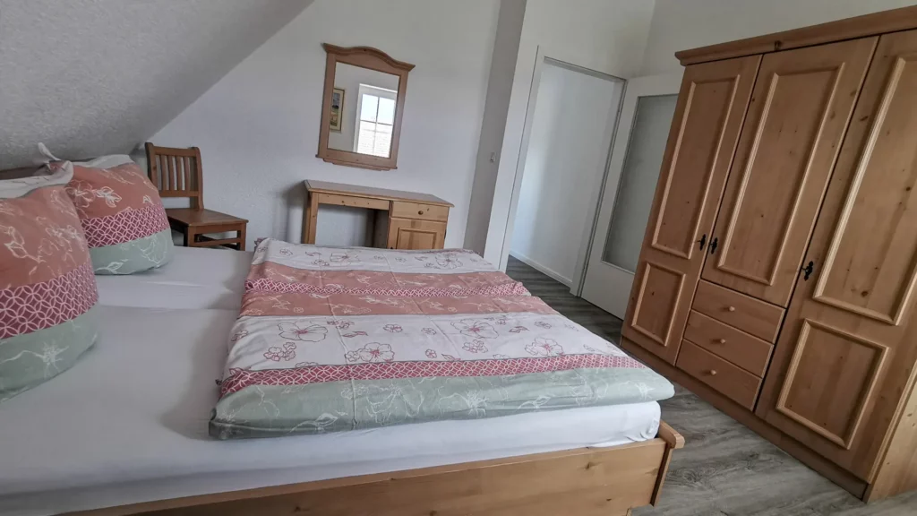 Komfortables Schlafzimmer der Ferienwohnung Jacqueline mit großem Ehebett, Schreibtisch, Spiegel, Stuhl, großem dreitürigem Kleiderschrank, schicken Accessoires und großem Fenster.