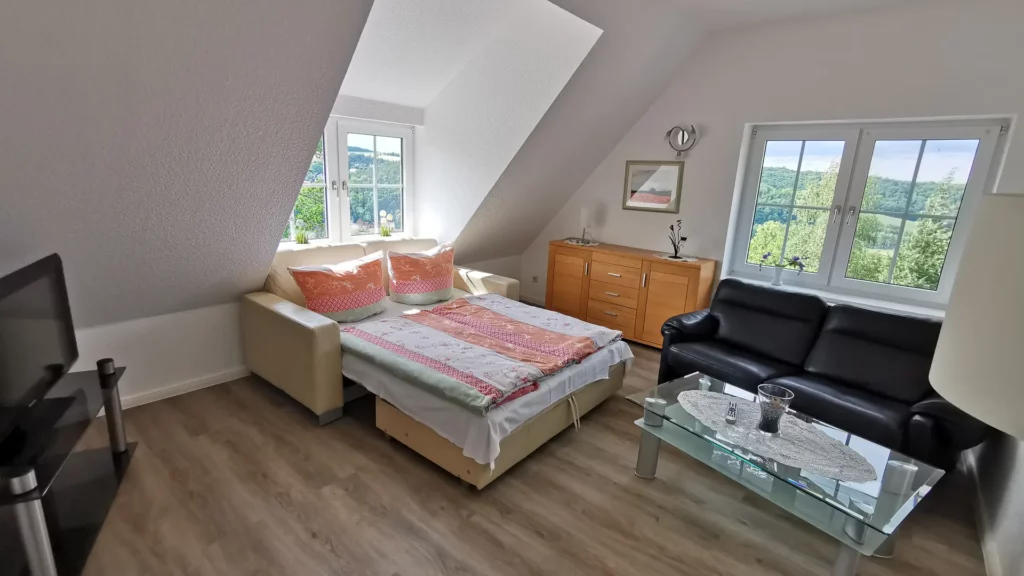 Wohnzimmer der Ferienwohnung Jacqueline mit ausziehbarer Couch mit Federkern, LCD-Fernseher und zwei großen Fenstern, die den Raum mit Licht durchfluten.