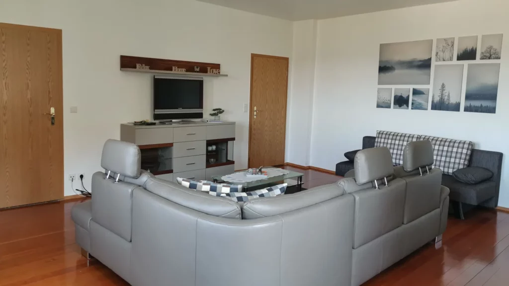 Wohnzimmer der Ferienwohnung Lisa mit großer lederbezogener Sitzlandschaft, zusätzlicher Schlafcouch, großer Sideboardanlage, LCD-Fernseher, Wandregalen und Lerchenparkett. Direkter Zugang zum Balkon mit Blick über den gesamten Ort.