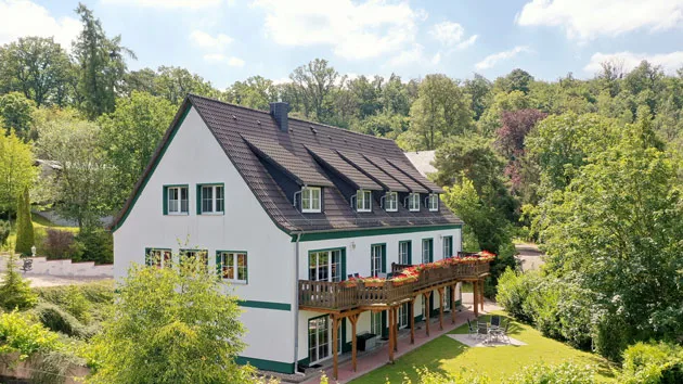 Luftaufnahme zeigt Ferienanlage mit Gebäuden, Freiflächen, Wald und Bergen im Hintergrund - mitten im Naturschutzgebiet.